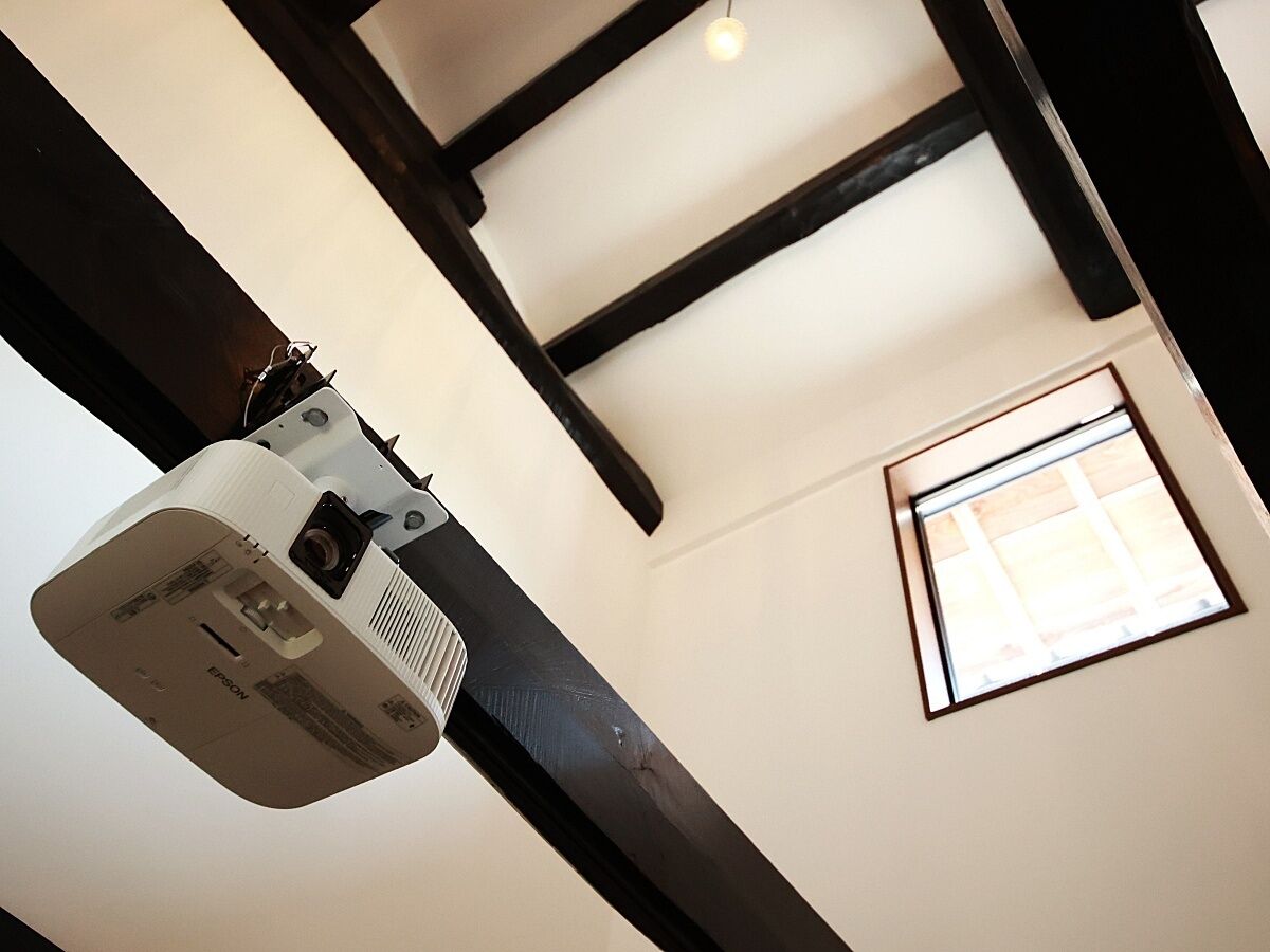 A projector mounted on a ceiling with exposed beams, a window with blinds, and a wall-mounted thermostat.