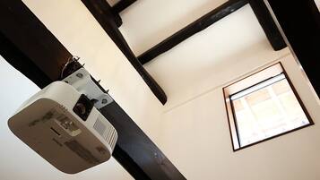 A projector mounted on a ceiling with exposed beams, a window with blinds, and a wall-mounted thermostat.