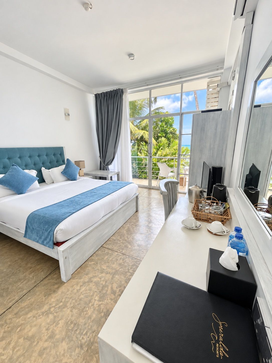 A hotel room with a bed, a desk, a chair, a TV, and a balcony with a view of palm trees.