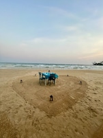Cenário de praia com mesa posta para dois, rodeada por lanternas, sob um céu claro.