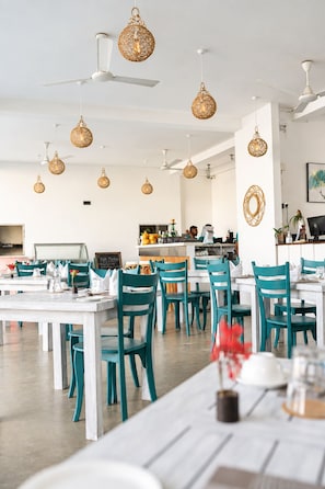Un comedor con varias mesas y sillas, ventiladores de techo e lámparas colgantes.