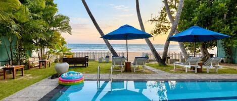 A swimming pool with clear blue tiles, surrounded by palm trees and lounge chairs, with a view of the ocean.