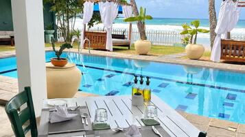 Mesa de comedor para dos con vista a la piscina, asientos exteriores y una valla blanca al fondo.