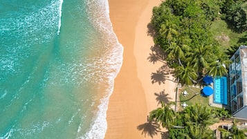 An aerial view of a beach with a pool area, palm trees, and a building with a blue roof.