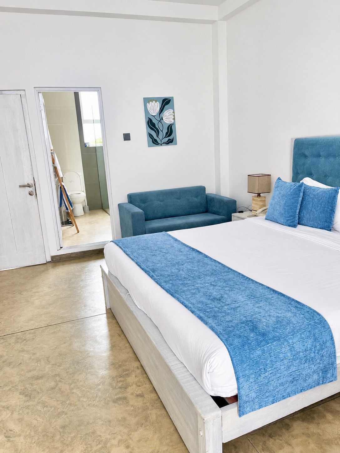 A bedroom with a large bed, a blue sofa, a painting on the wall, and a bathroom visible through an open door.