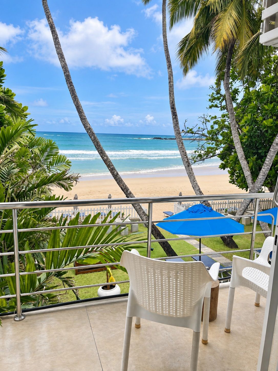 A balcony with white wicker chairs, a blue umbrella, and a view of the beach and ocean.