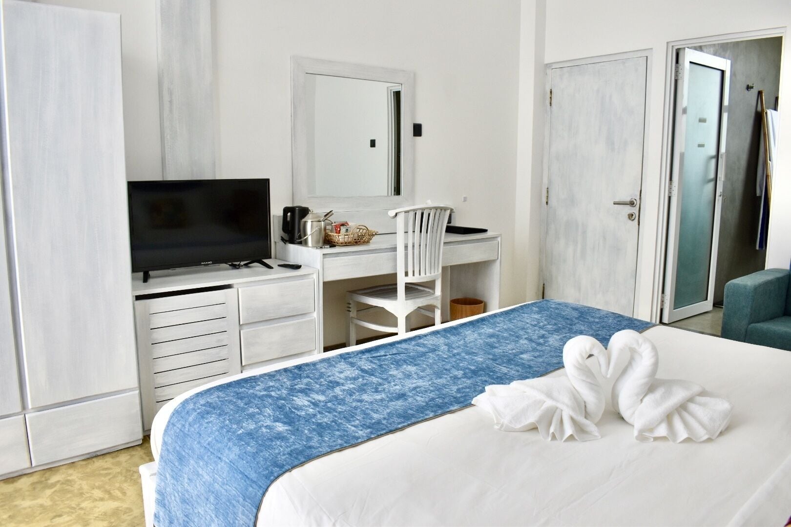 Quarto de hotel com cama, escrivaninha com cadeira, televisão e guarda-roupa.