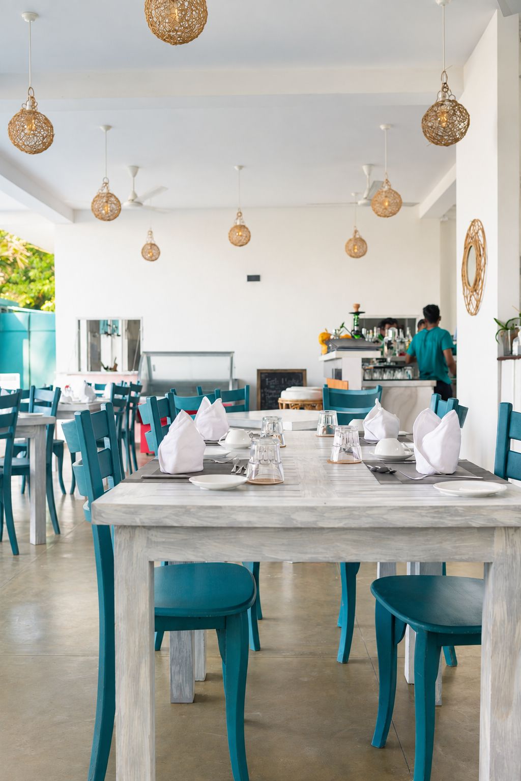 Un coin repas avec des chaises de couleur turquoise et une table blanche dressée pour quatre personnes, des suspensions en osier au-dessus, et un espace cuisine en arrière-plan.