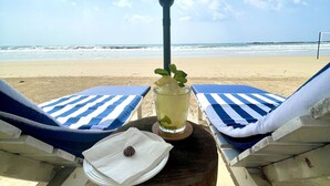 Blick auf einen Strand mit zwei Liegestühlen unter einem Sonnensegel, einem Glas Eistee mit Minze und einem Holzschemel.