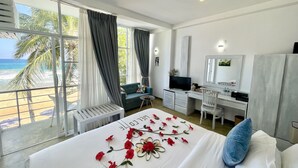 Ein Hotelzimmer mit Bett, Schreibtisch, Stuhl, Fernseher und einem Balkon mit Blick auf den Strand und Palmen.