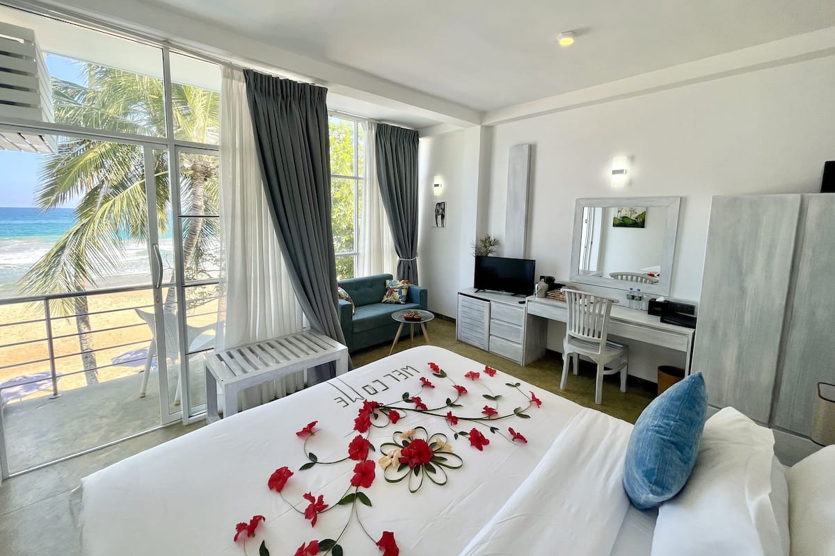 A hotel room with a bed, a desk, a chair, a TV, and a balcony with a view of the beach and palm trees.