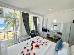 Ein Hotelzimmer mit Bett, Schreibtisch, Stuhl, Fernseher und einem Balkon mit Blick auf den Strand und Palmen.