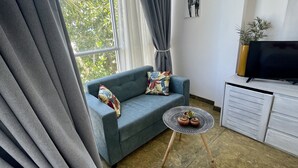 Ein modernes Wohnzimmer mit einem grauen Sofa, einem kleinen runden Tisch mit zwei Topfpflanzen, einem Fernseher auf einem weißen Schrank und einem großen Fenster mit grauen Vorhängen.