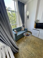 A modern living room with a grey sofa, a small round table with two potted plants, a TV on a white cabinet, and a large window with grey curtains.