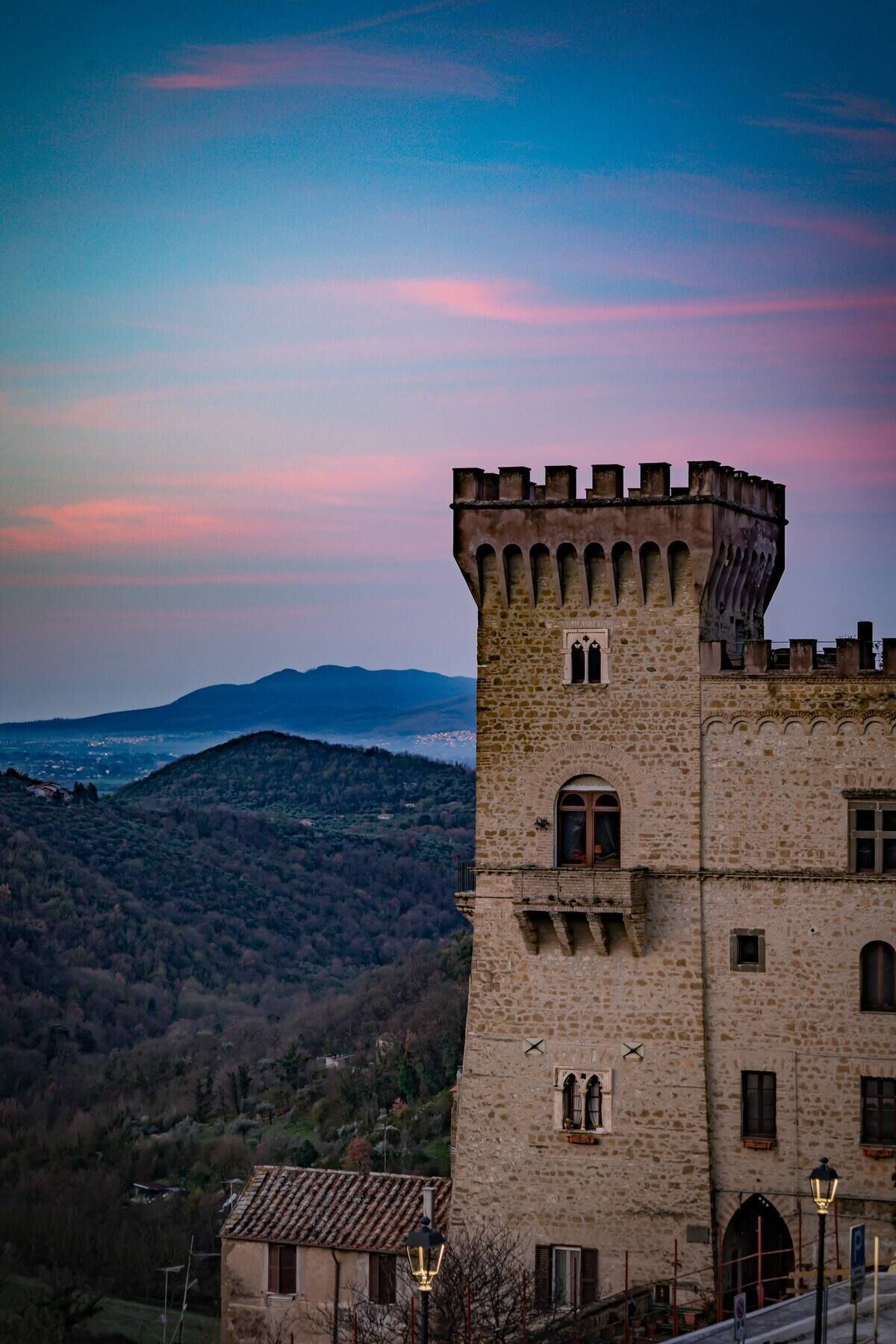 A stone tower with arched windows and a battlement, overlooking a valley with distant mountains and a town.