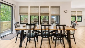 Un espace repas avec une table en bois et des chaises noires, de grandes fenêtres équipées de stores et une vue sur la verdure à l’extérieur.
