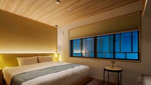 A hotel room with a large bed, a bedside table with a lamp, a window with blinds, and a small round table with a vase.