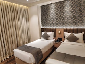 Habitación de hotel con dos camas, un ventanal con cortinas, un papel tapiz estampado y una mesita de noche con teléfono y lámpara.