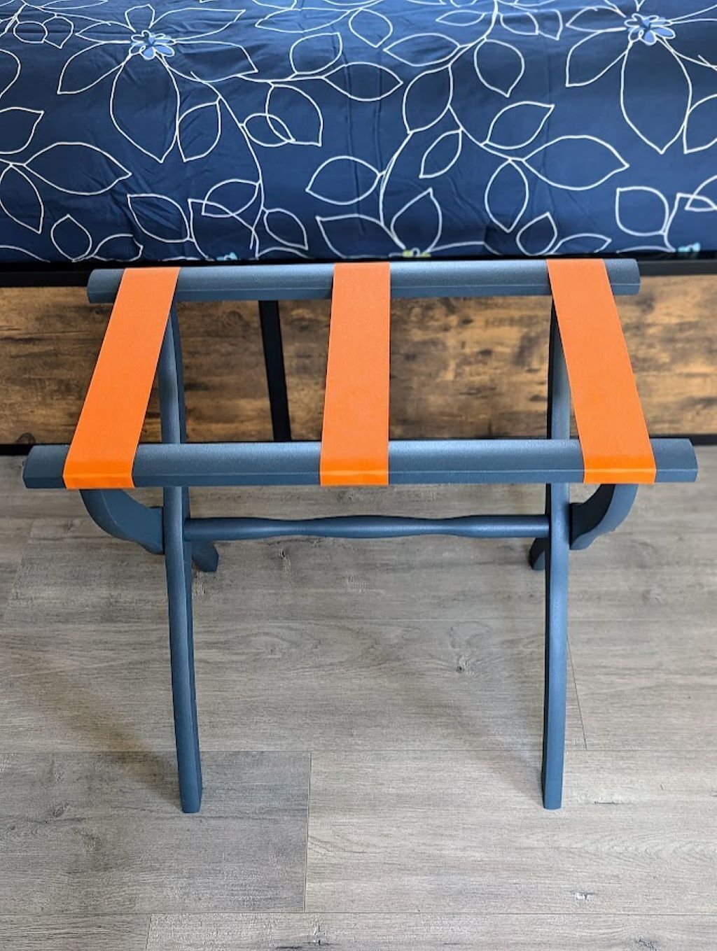 A bench with orange straps and a dark blue frame, placed on a wooden floor with a floral-patterned bedspread in the background.