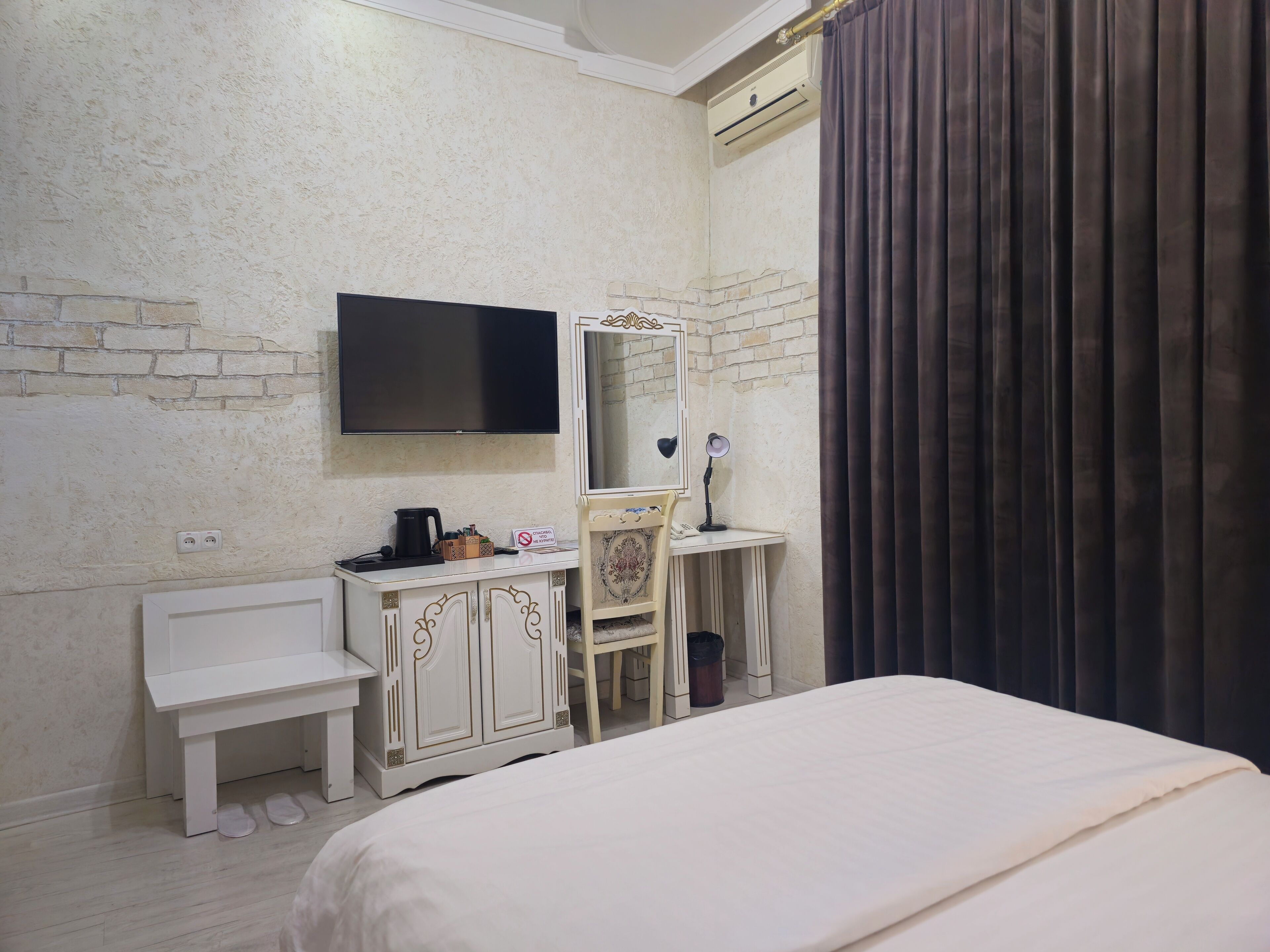 A hotel room with a bed, a desk with a chair, a television mounted on the wall, and a window with curtains.