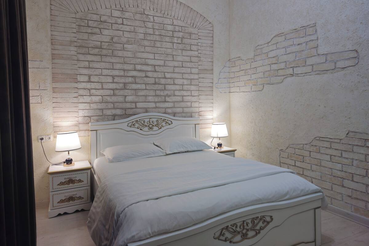 Un dormitorio con pared de ladrillo visto, una cama blanca con cabecero decorativo, dos mesitas de noche con lámparas y un mueble auxiliar con cajones.