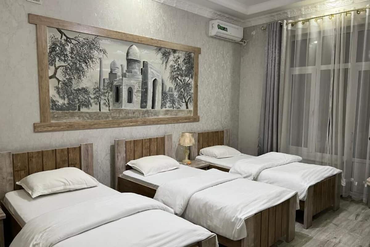 Habitación de hotel con cuatro camas individuales, un cuadro grande enmarcado en la pared, un equipo de aire acondicionado y una ventana con cortinas traslúcidas.