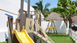 A pirate-themed play structure with a slide and climbing area, set in a grassy yard with a white fence and palm trees in the background. - Vacation getaway with beach access & balcony (CancÃºn)