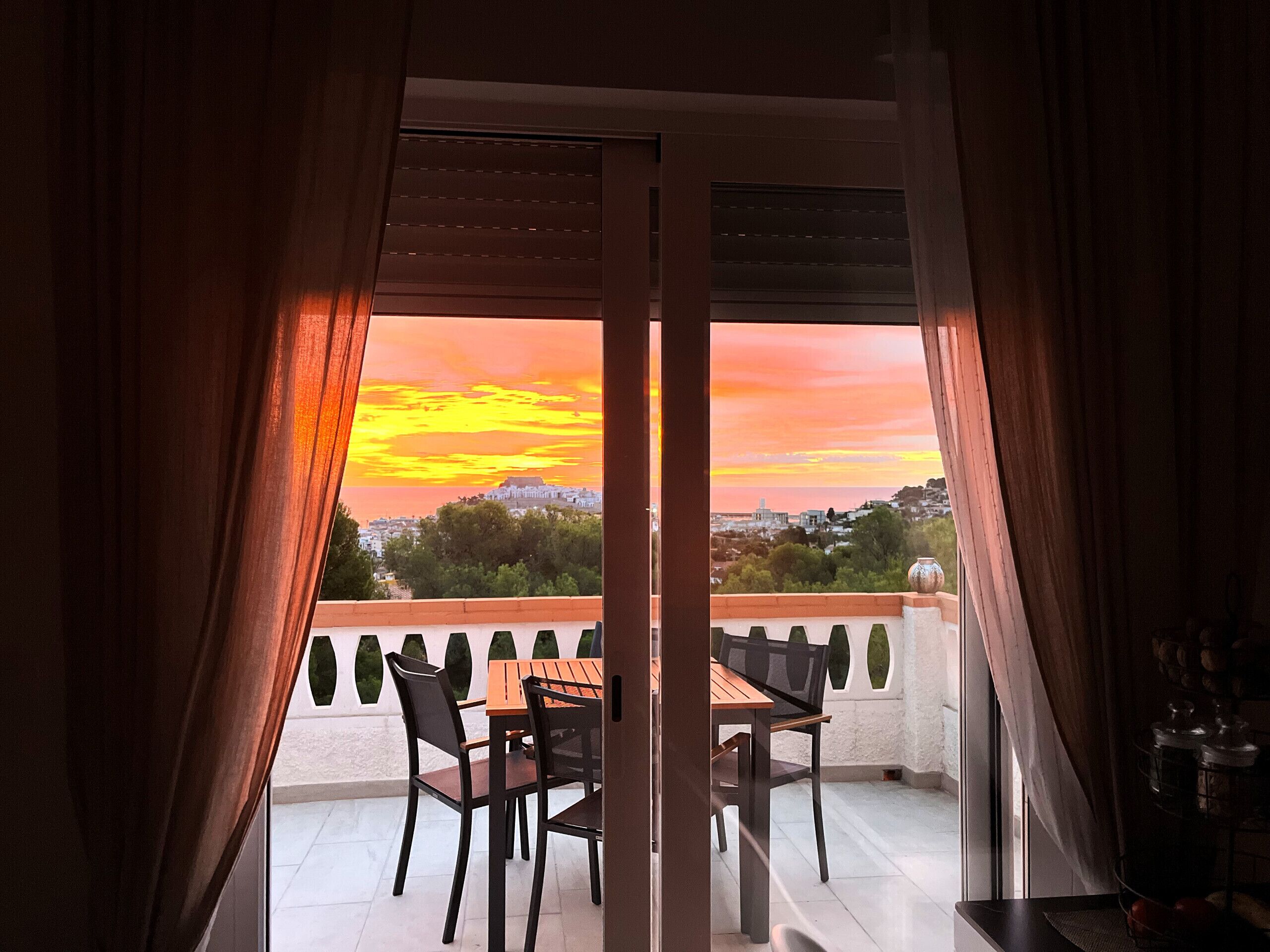 Un balcón con mesa y sillas, con vista a un paisaje urbano al atardecer.