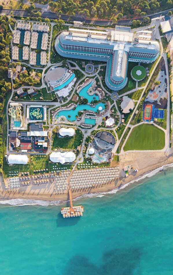 An aerial view of a resort with multiple swimming pools, a beach area, and a large building with a curved roof. - Ethno Belek (Serik)