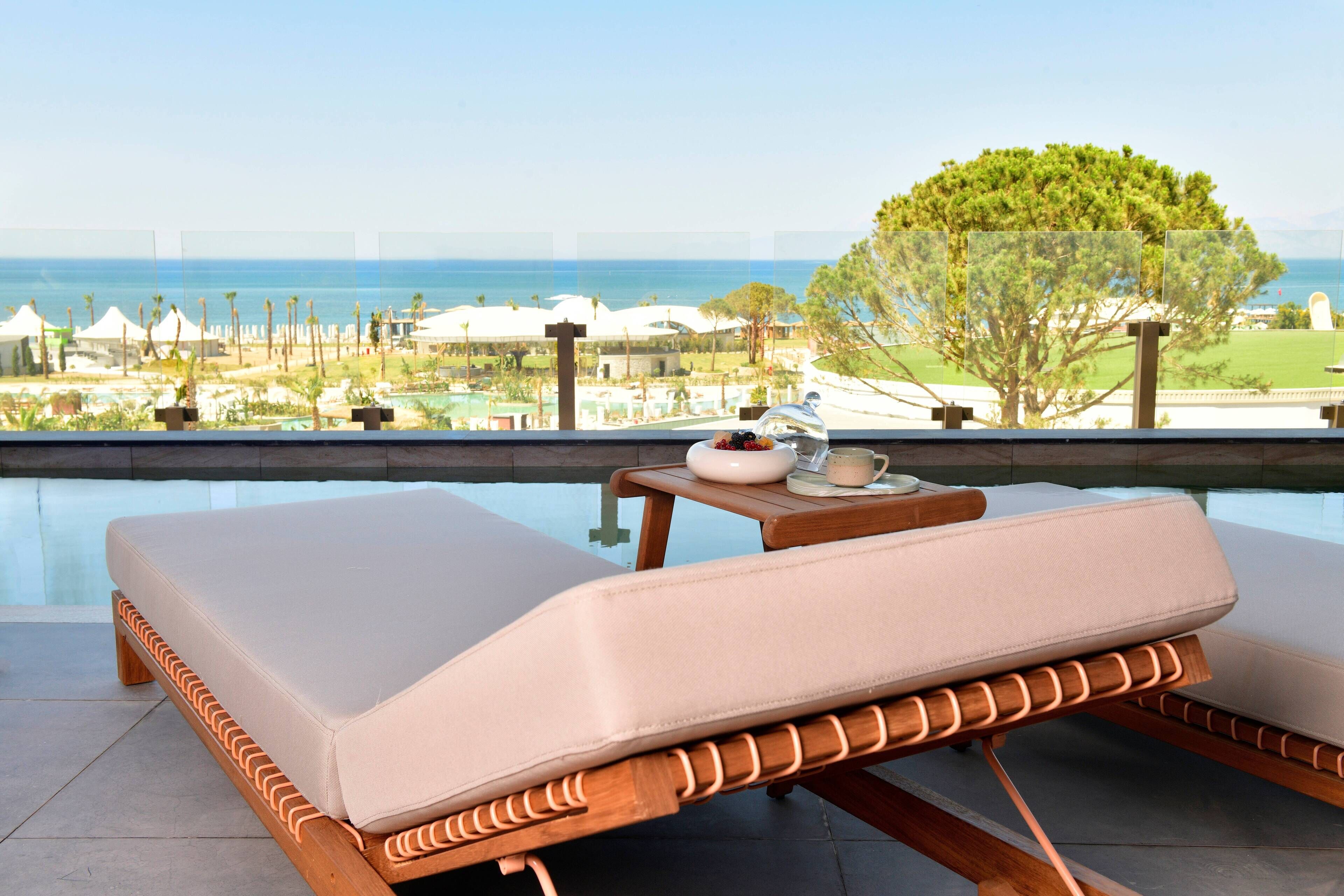 A lounge chair with a cushion, a small table with a teapot and cup, and a view of a pool and resort area.