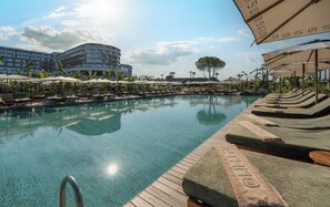 A clear pool with lounge chairs and umbrellas, surrounded by palm trees and a modern building in the background. - Ethno Belek (Serik)