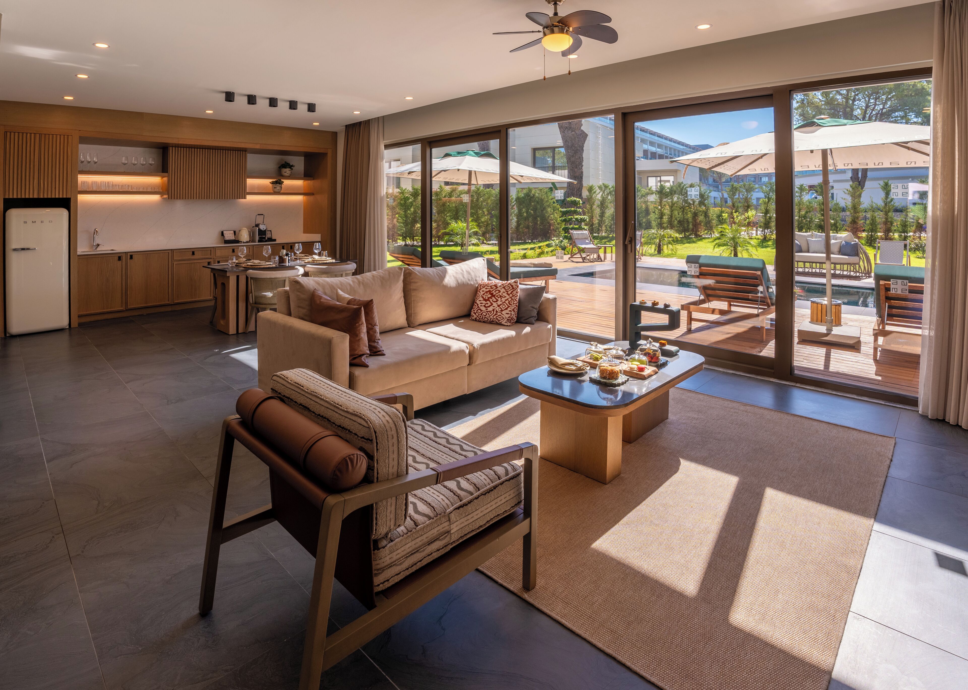 Amplia sala de estar con sofá, sillón y mesa de centro, cocina con alacenas de madera y un gran ventanal corredero que da a un patio exterior con sombrillas.