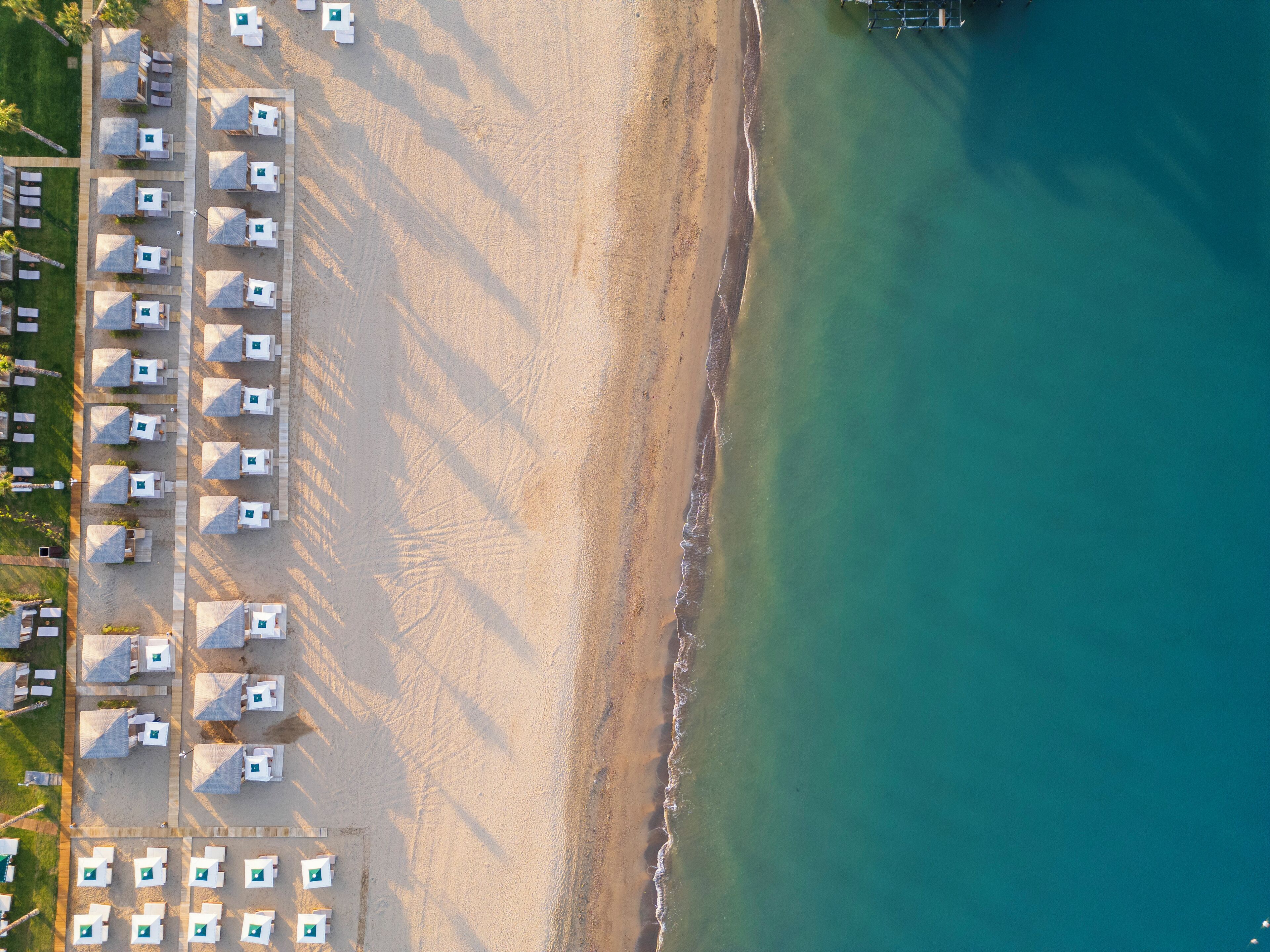 Vue aérienne d’un complexe hôtelier en bord de mer, avec des rangées de chambres donnant sur la plage, une plage de sable et une mer d’un bleu limpide.