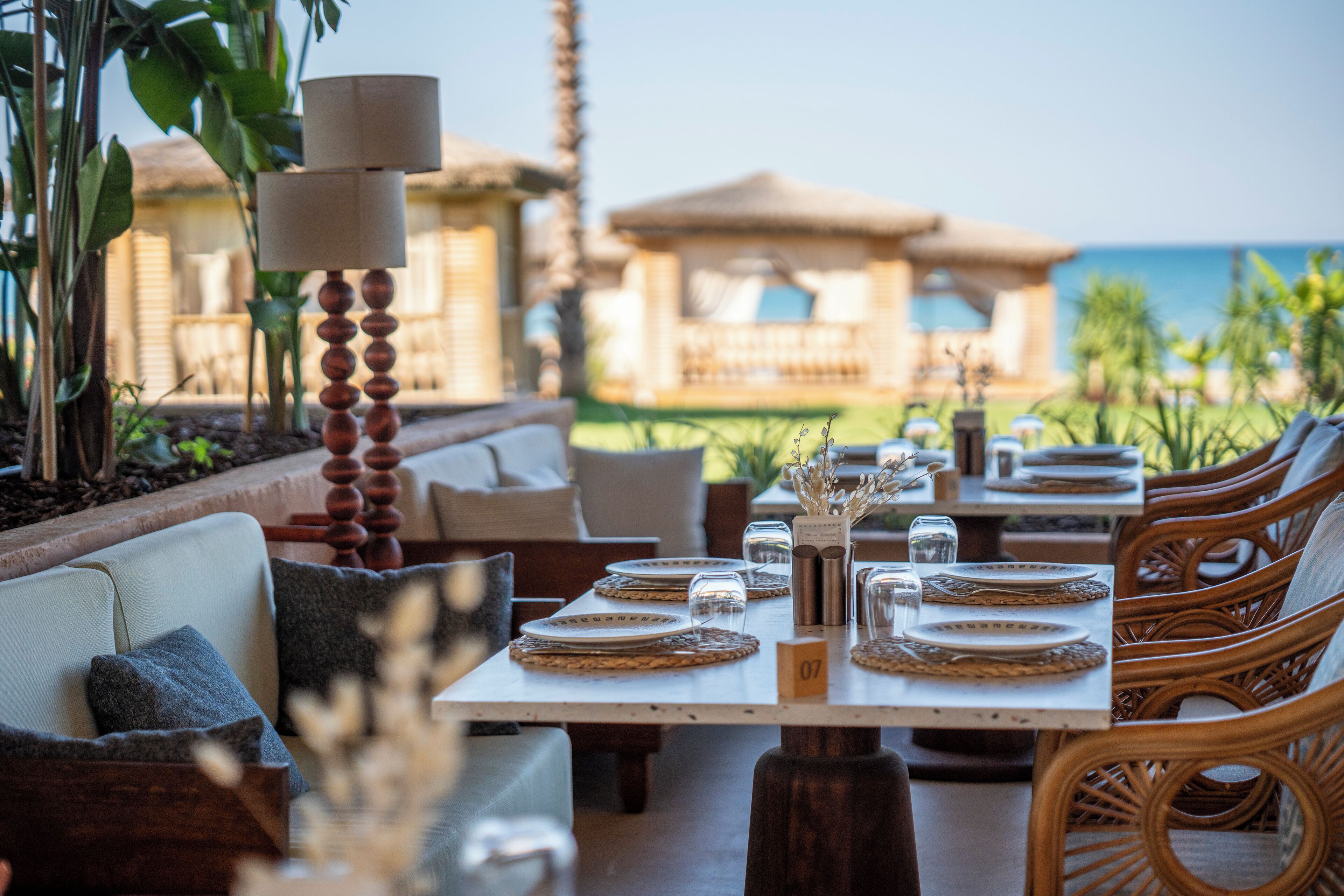 An outdoor dining area with wicker furniture, round tables, and a view of the ocean.