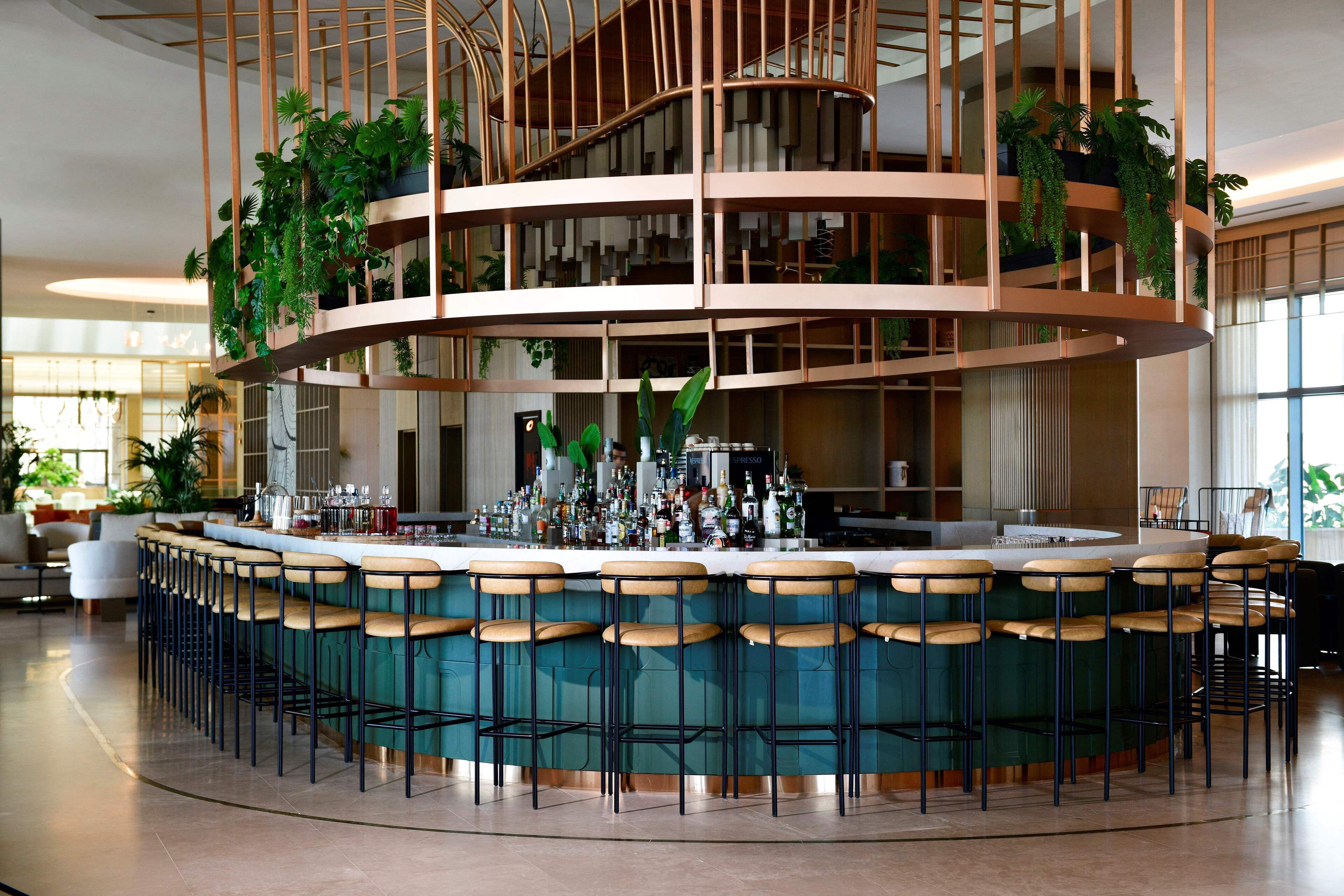 Zona de bar moderna con varios taburetes y una barra central, rodeada por un espacio de estar circular y una estructura de jardín vertical.