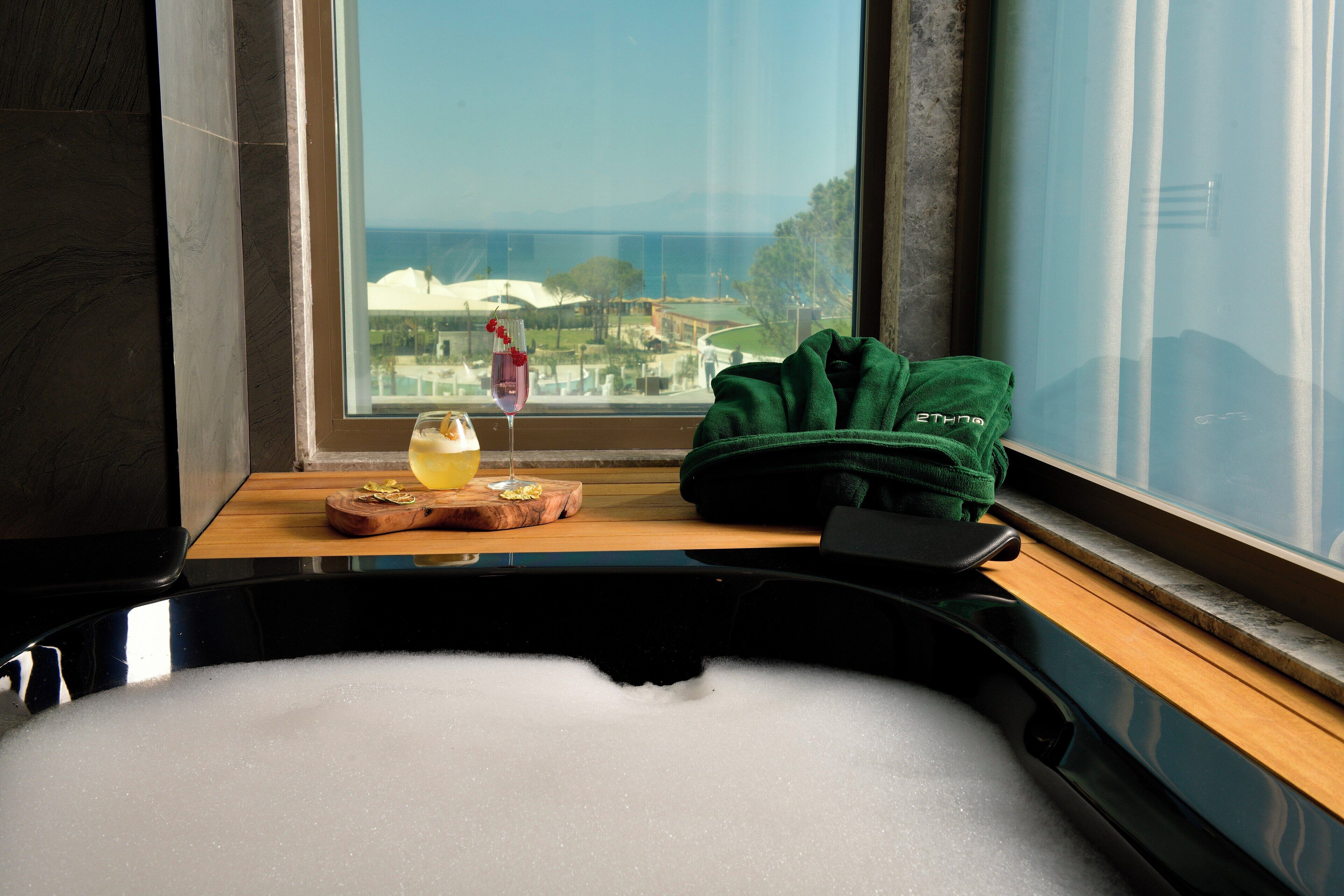 A bathtub with a view of the ocean, a towel, and a glass of a pink drink on a wooden ledge.