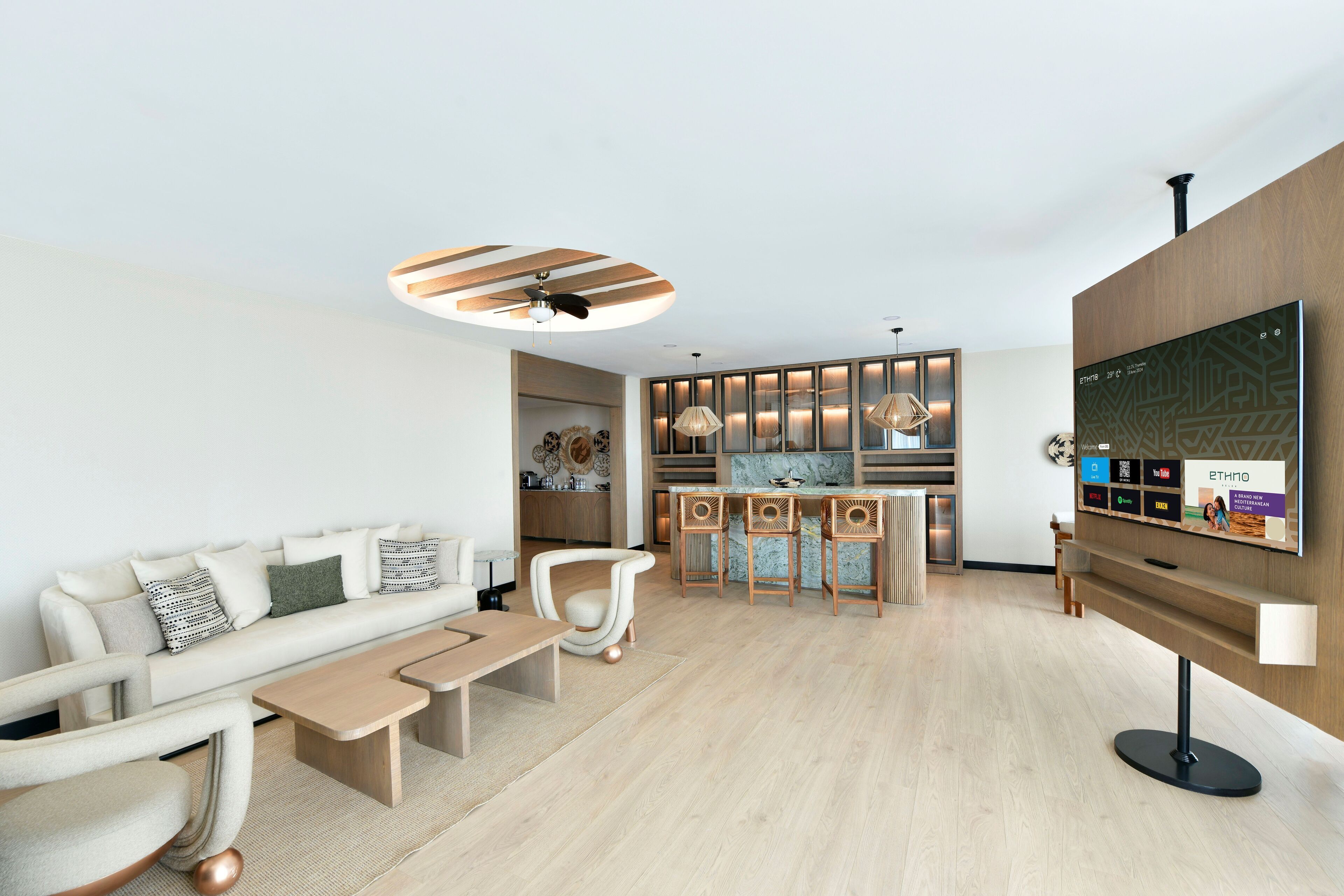 Una sala de estar moderna con sofá, mesa de centro y sillones. Se observa una zona de cocina con taburetes altos y lámparas colgantes. Un televisor de pantalla plana está fijado a la pared.