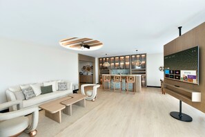 A modern living room with a sofa, coffee table, and chairs. There is a kitchen area with bar stools and pendant lights. A flat-screen TV is mounted on the wall.