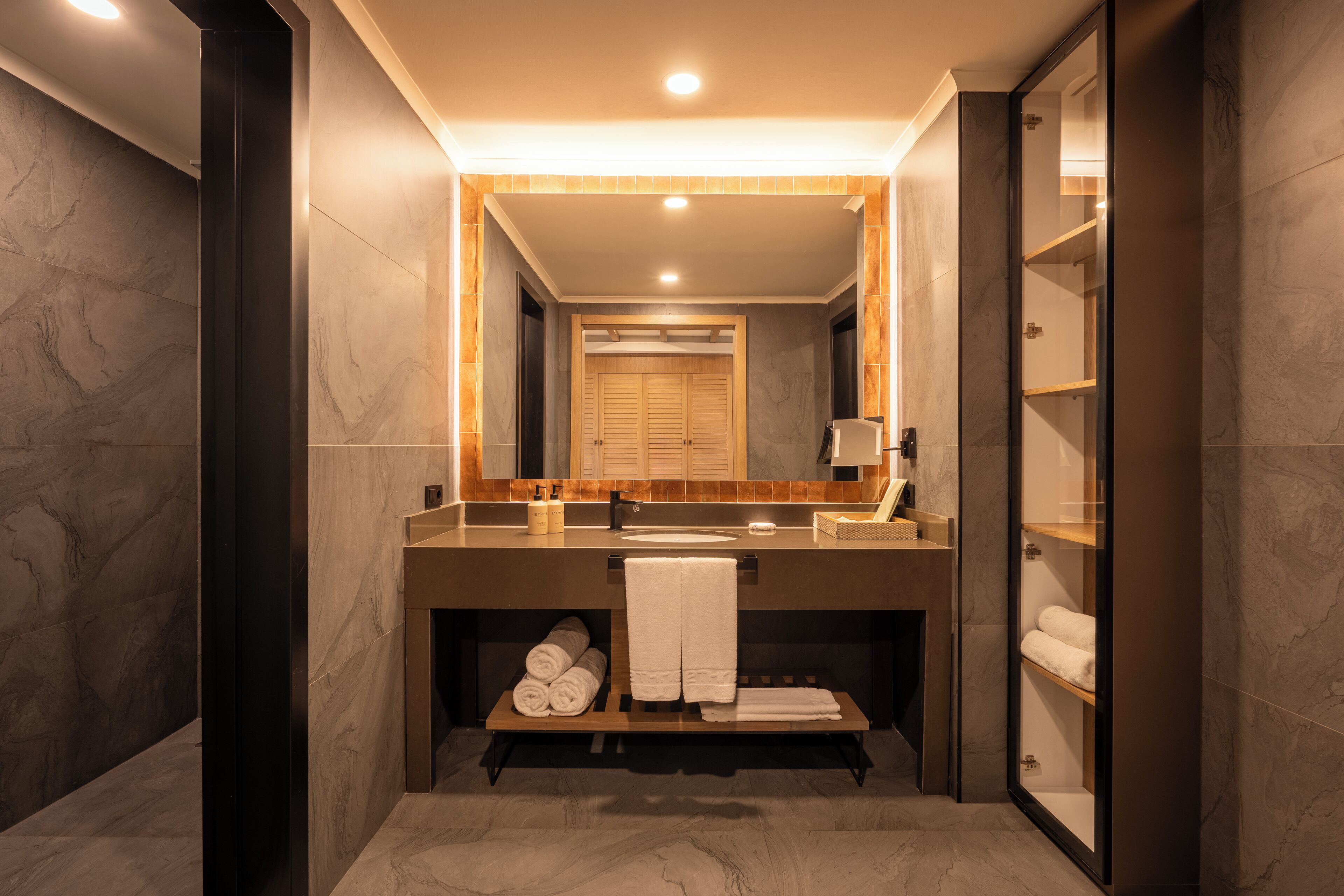 A modern bathroom with a large mirror, dark marble walls, a wooden vanity, and a built-in shelf with towels.