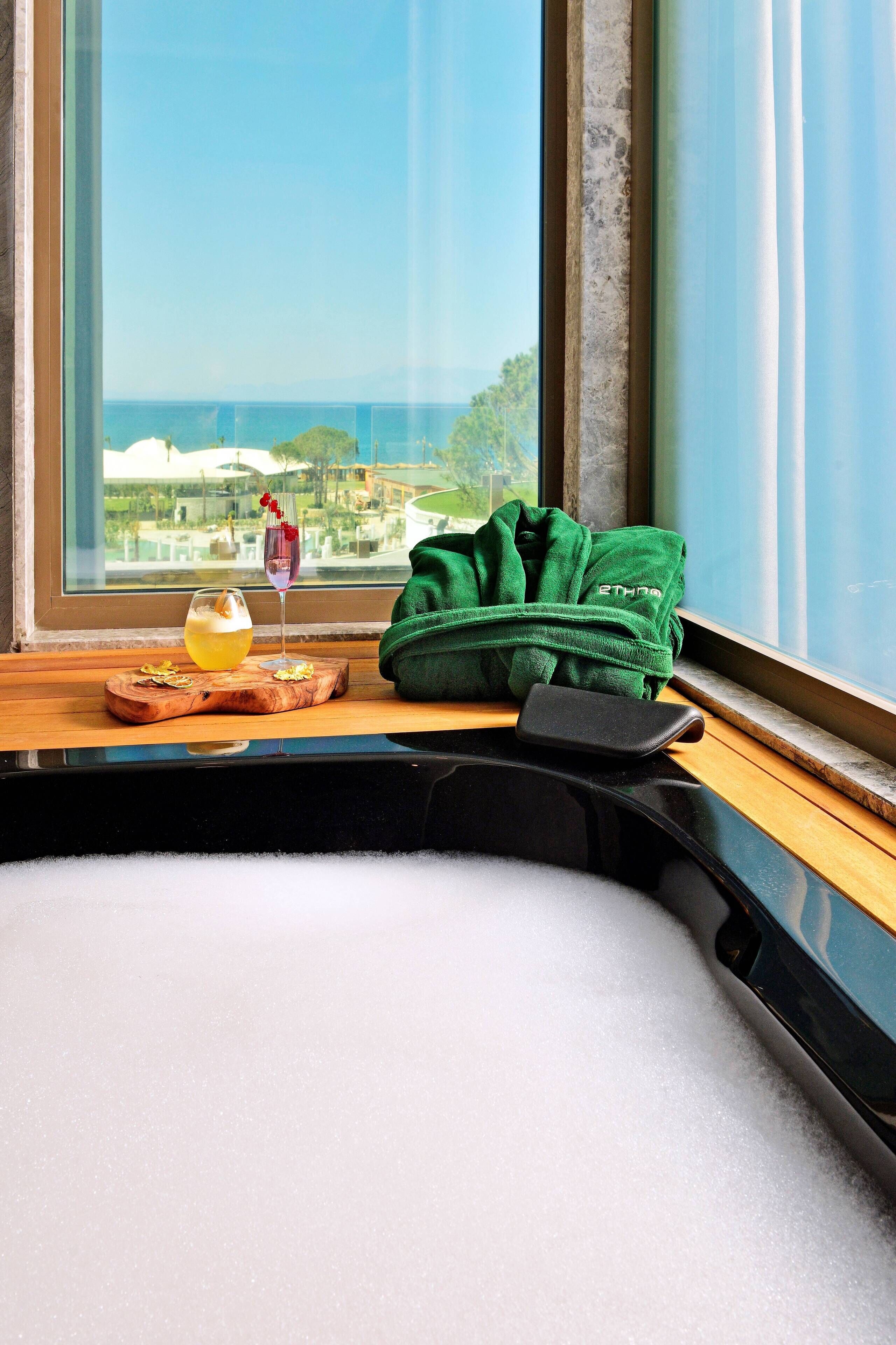 A window seat with a view of the sea, a green towel, a glass of drink, and a wooden tray.