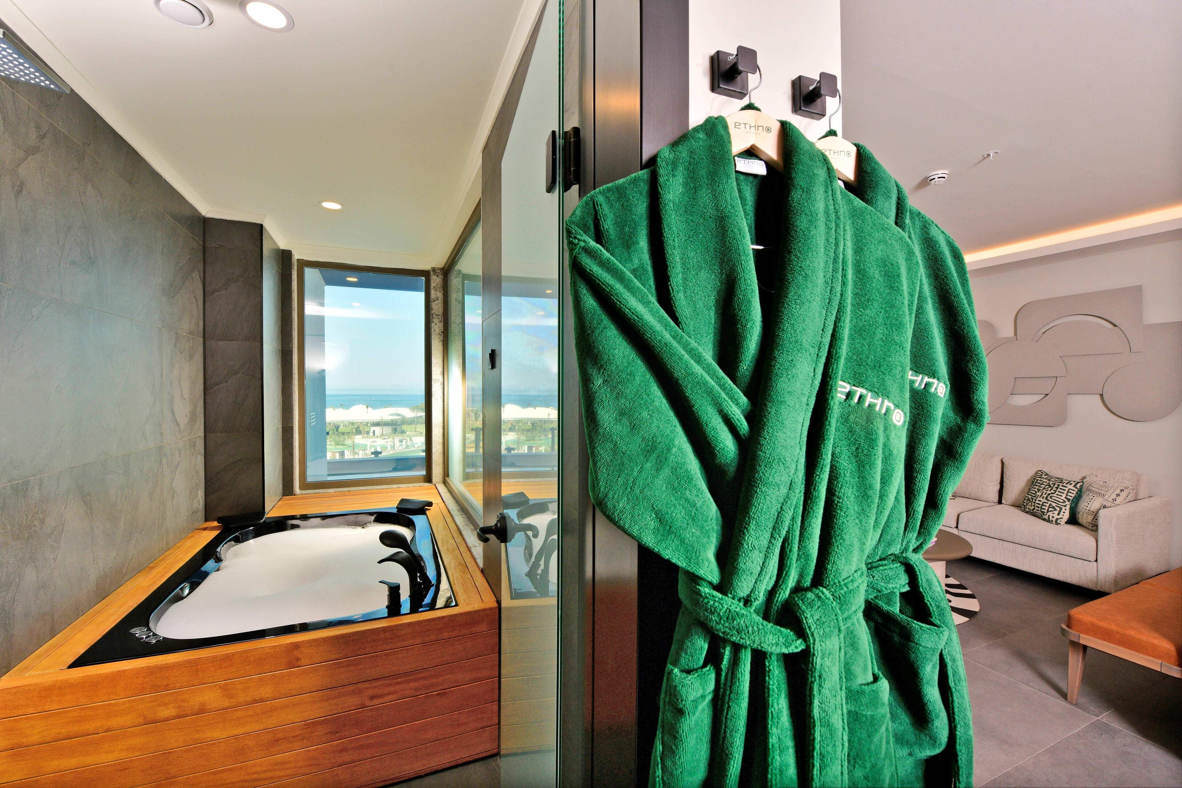 A modern bathroom with a wooden bathtub, a green robe hanging on a rack, and a view of the city through large windows.