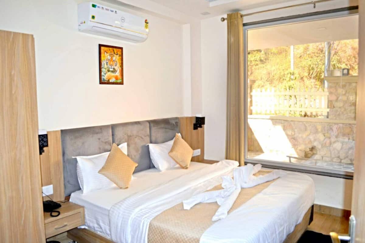 A hotel room with a large bed, a bedside table, an air conditioning unit, and a window with a view of trees.