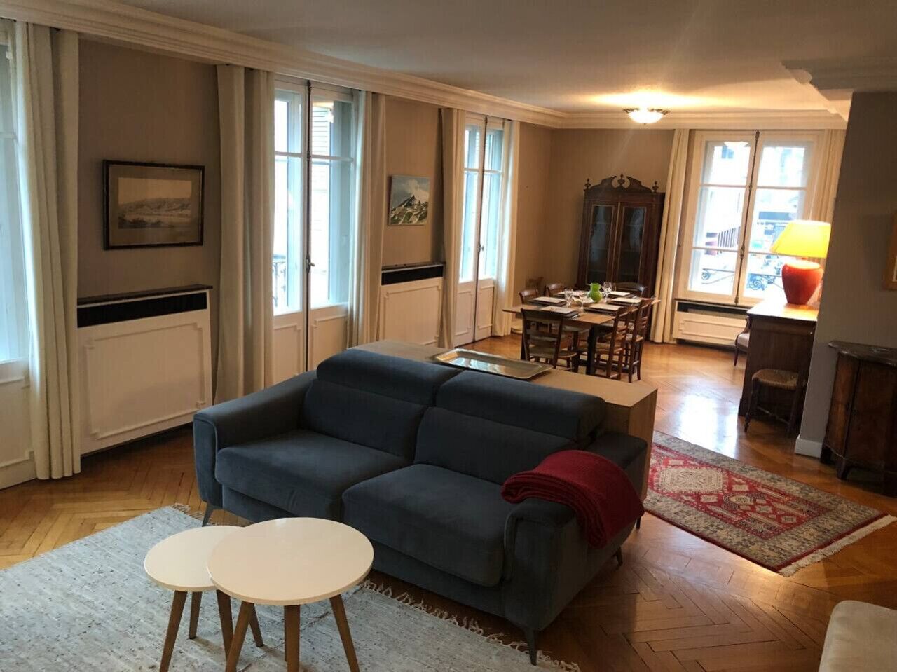 Una sala de estar con un sofá azul, una mesita redonda y una alfombra roja. Se observa un comedor con una mesa y sillas al fondo.