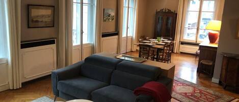 Uma sala de estar com um sofá azul, uma mesa redonda pequena e um tapete vermelho. É possível ver, ao fundo, uma área de jantar com uma mesa e cadeiras.
