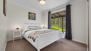 A bedroom with a white bed, a nightstand, a bedside lamp, a painting, and a window with curtains. - Ellendale - enjoy the serenity (Kurmond)