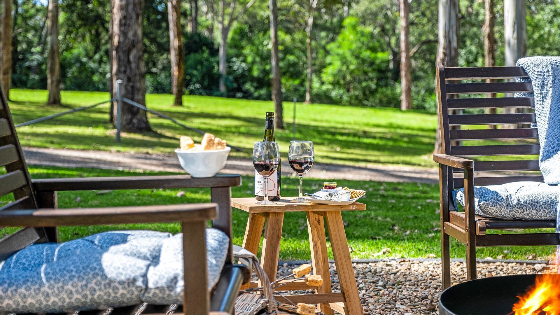 Ein Picknick mit einer Weinflasche, zwei Gläsern, einer Brotschale und einem Feuerkorb in einem Waldgebiet.