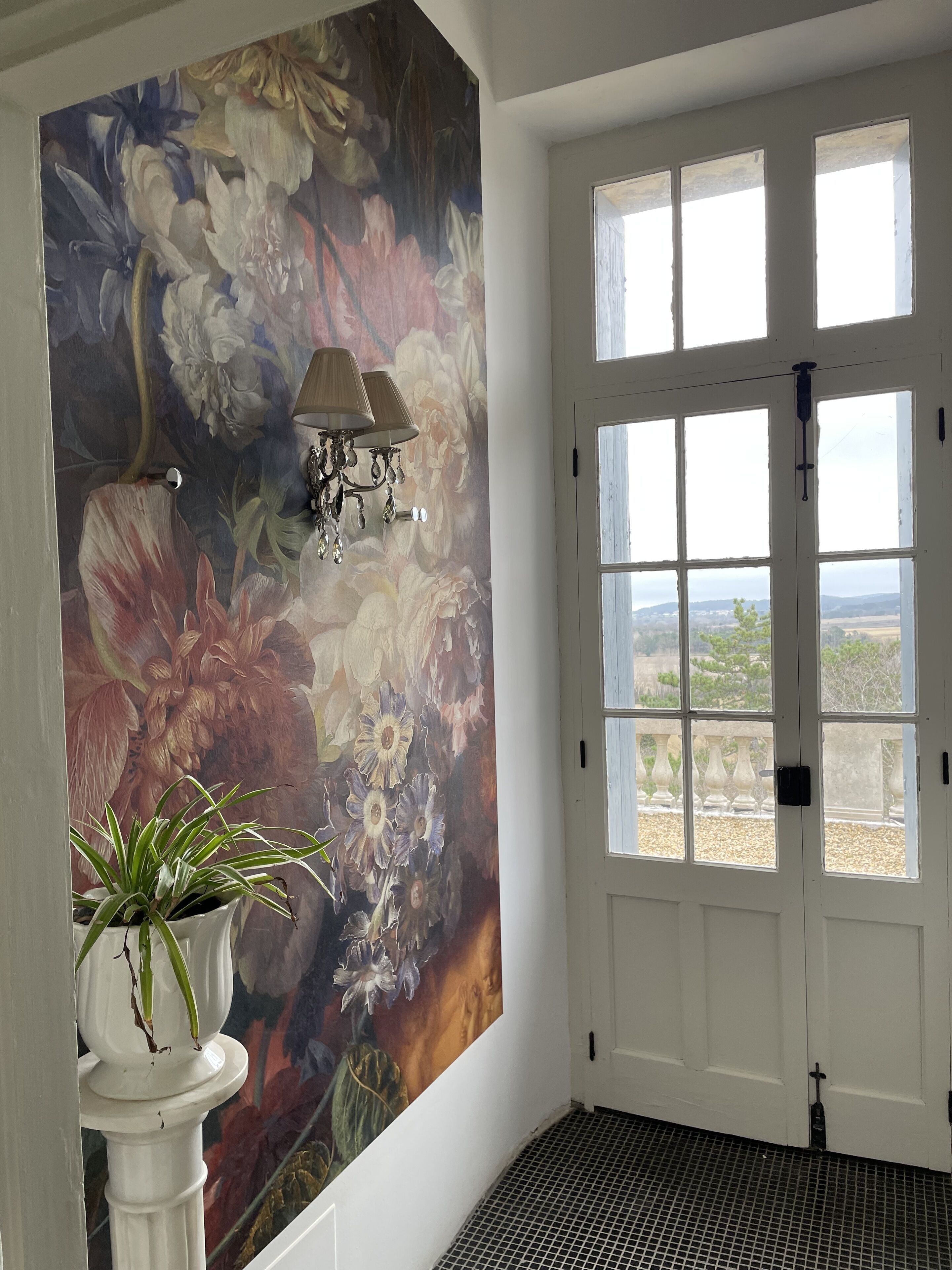 Une pièce décorée d’une grande fresque florale, avec une porte blanche dotée de plusieurs fenêtres et une plante en pot.