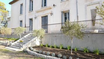 Un bâtiment à deux étages, doté d’un escalier en pierre menant à l’entrée, et entouré d’un jardin planté de jeunes arbres et d’arbustes.