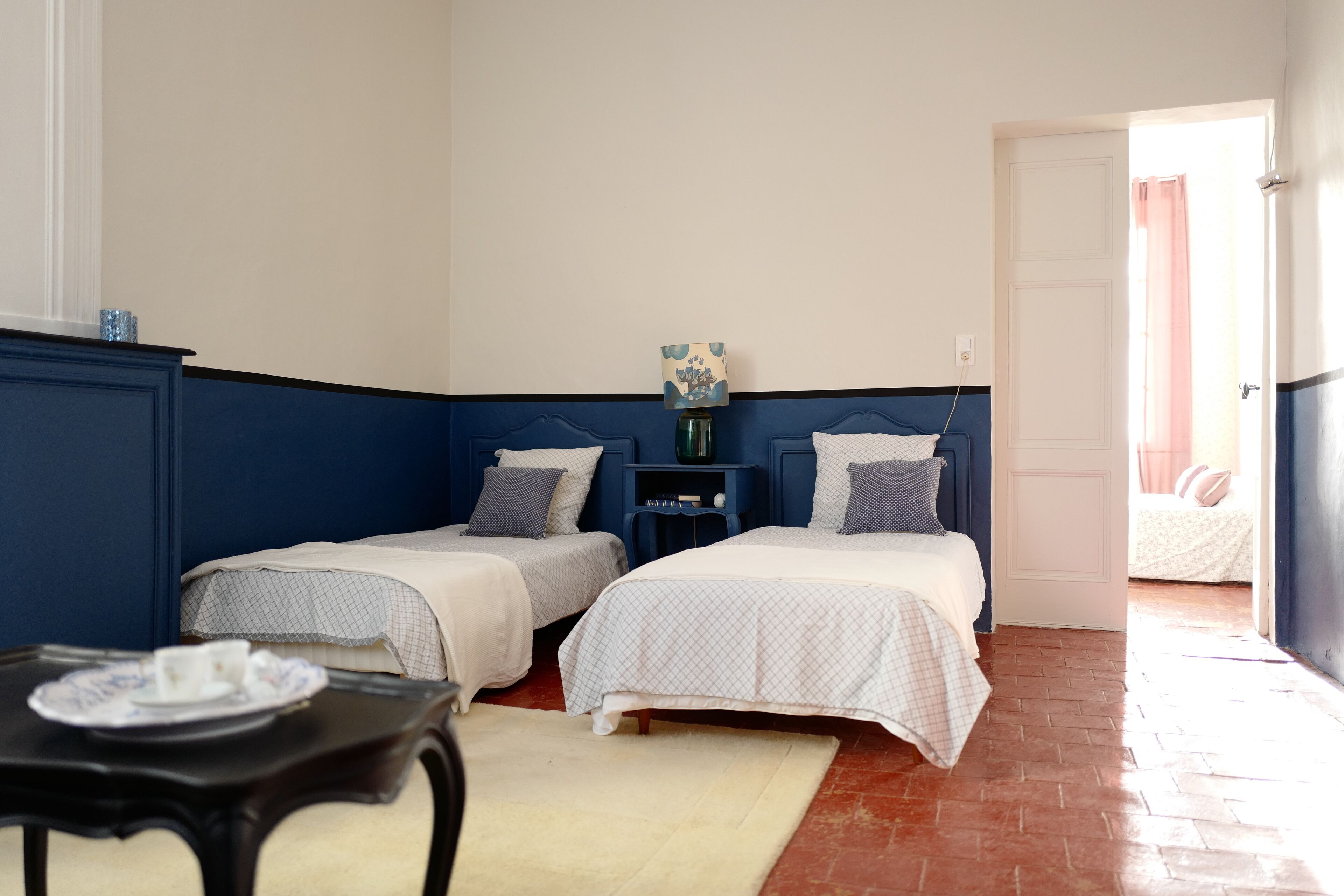A room with two beds, a small table with a cup and saucer, a nightstand with a lamp, and a door leading to another room.