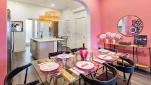 A dining area with a glass table set for six, a kitchen in the background, and a pink accent wall. - Curated Bachelorette Retreat w/ Balcony, Glam Room, Roof | Close to Downtown! (Nashville)